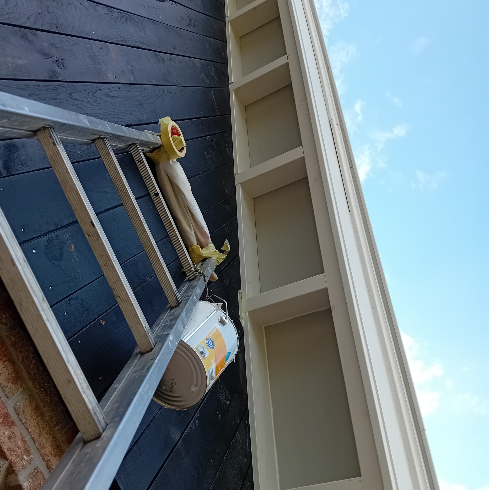 Metalowa drabina oparta o pomalowaną na ciemno drewnianą ścianę i jasną elewację, z zawieszoną puszką farby i taśmą malarską, widok z dołu na błękitne niebo.