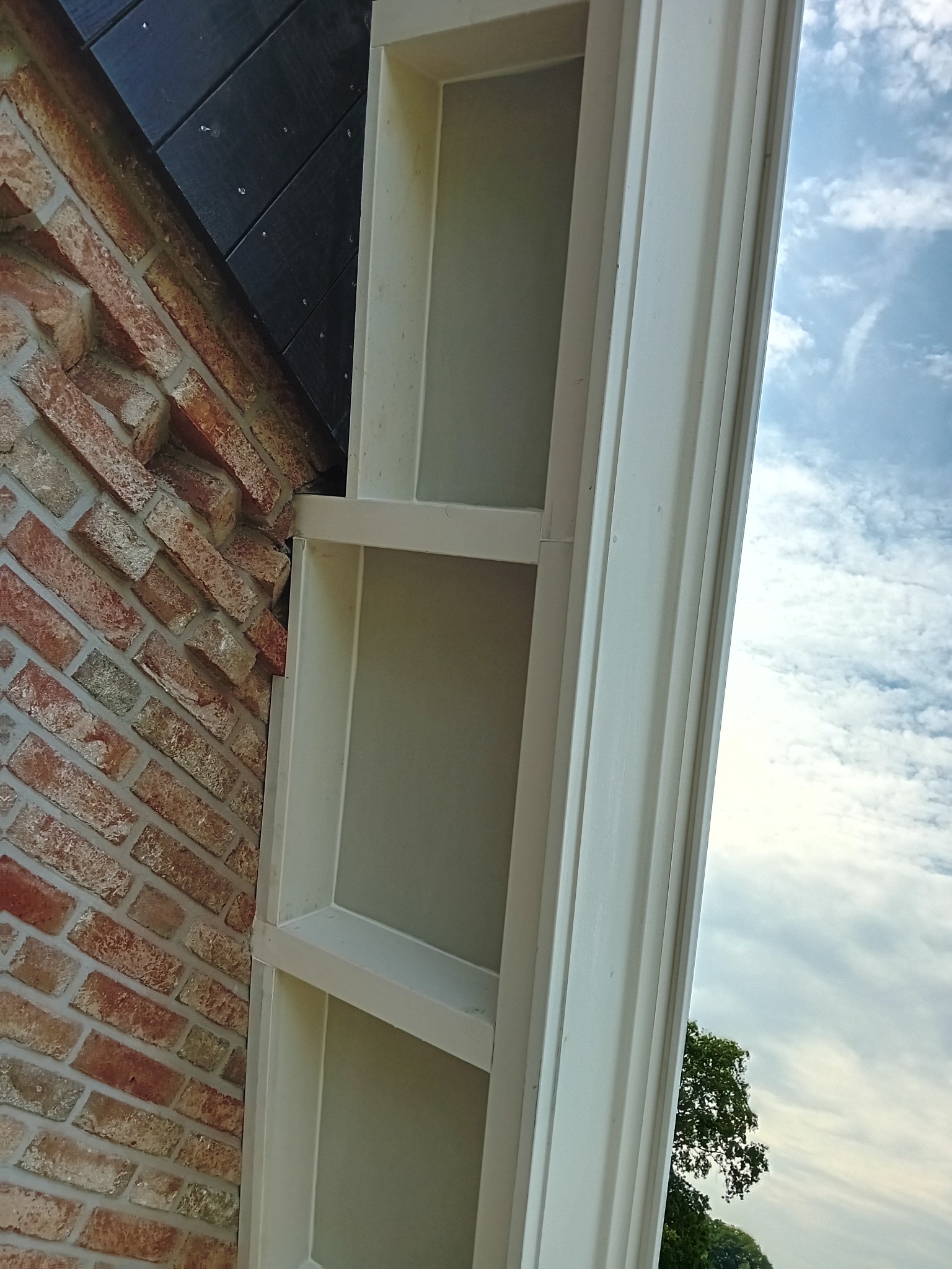 Fragment elewacji budynku z cegły klinkierowej, z widocznym białym, drewnianym elementem dekoracyjnym przypominającym pilaster, na tle błękitnego nieba z chmurami.