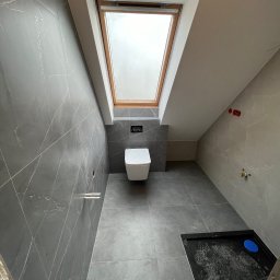 Maks handzlik - Wnętrze łazienki na poddaszu z szarymi płytkami imitującymi marmur, wiszącą toaletą, oknem dachowym i brodzikiem w trakcie montażu.