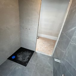 Maks handzlik - Wnętrze łazienki w trakcie wykańczania, widoczny czarny brodzik z niebieskim odpływem, szare płytki imitujące marmur na ścianach i podłodze, surowy otwór drzwiowy z widoczną podłogą z płyt OSB...