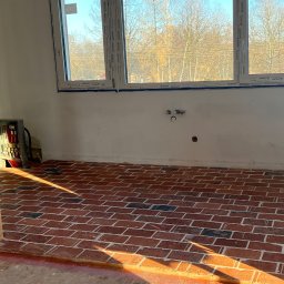 Maks handzlik - Pomieszczenie w trakcie wykańczania z ceglaną podłogą w jodełkę, widoczne białe ściany z instalacją hydrauliczną i trzy okna z widokiem na drzewa, słoneczne światło pada na podłogę.