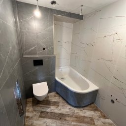 Maks handzlik - Wykończona łazienka z szarymi i białymi płytkami imitującymi marmur, drewnopodobną podłogą, białą wanną z szarym obramowaniem i podwieszaną toaletą, widoczne niedokończone punkty oświetleniowe.