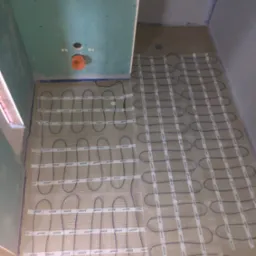 Montaż ogrzewania podłogowego w łazience, widoczne kable grzewcze ułożone na siatce, surowe ściany z płyt gipsowo-kartonowych, przygotowanie do wylewki.
