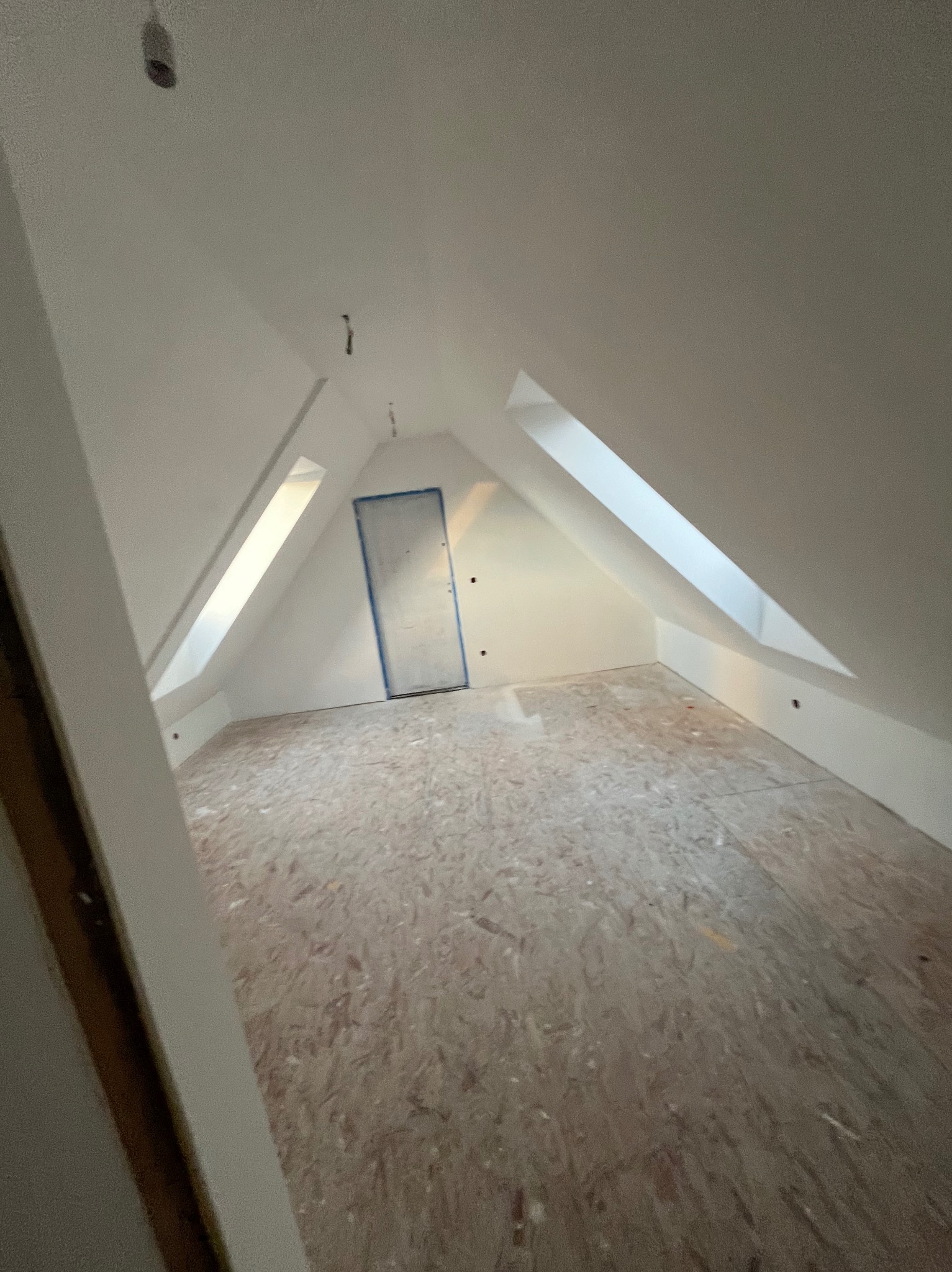 Poddasze z oknami dachowymi, przygotowane do wykończenia, z surową podłogą z płyt OSB i zabezpieczonymi taśmą malarską drzwiami.