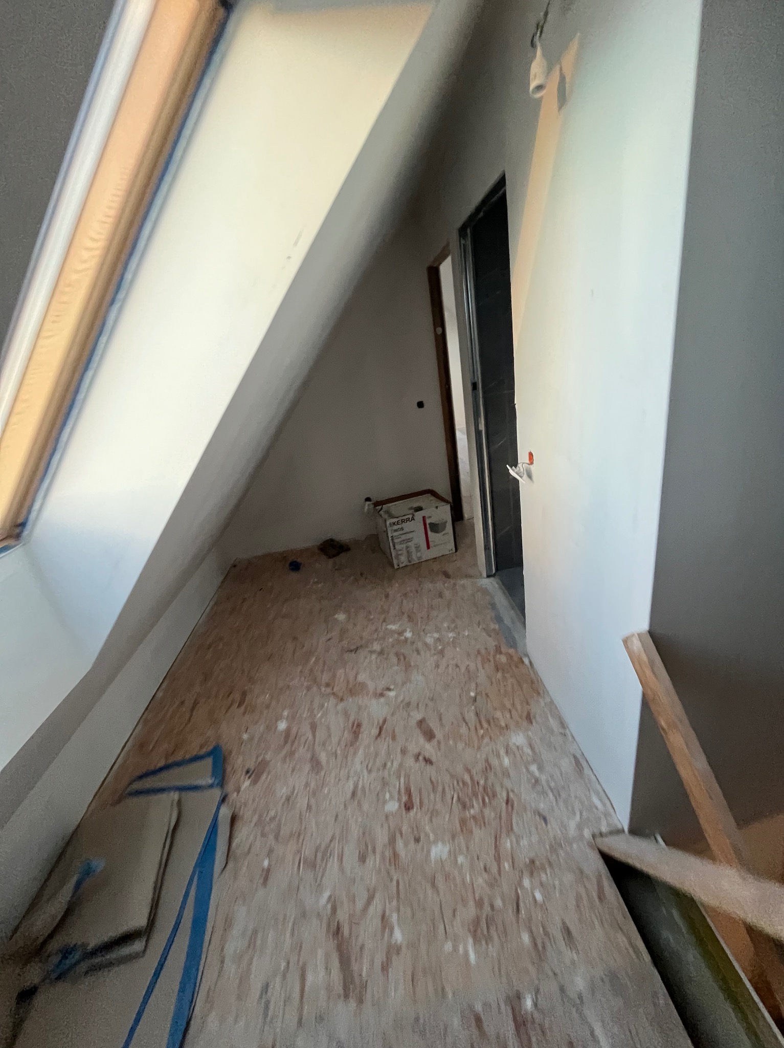 Poddasze w trakcie adaptacji: surowa podłoga z płyty OSB, okno dachowe z niebieską taśmą malarską, białe ściany w szarym pomieszczeniu, kartonowe pudło w głębi.