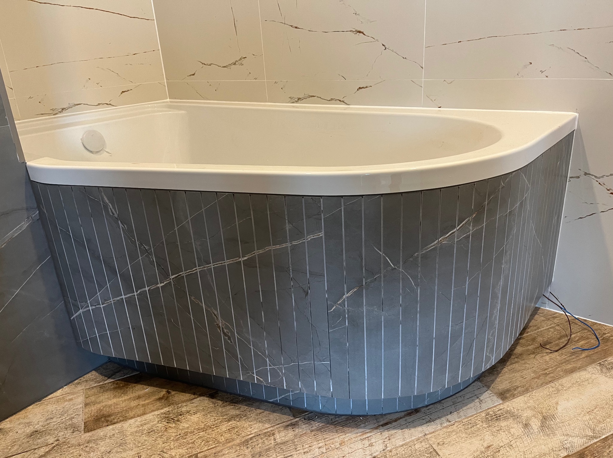 Biała wanna narożna z szarą obudową imitującą marmur, wykończona pionowymi listwami, na tle płytek imitujących marmur i drewnopodobnej podłogi.