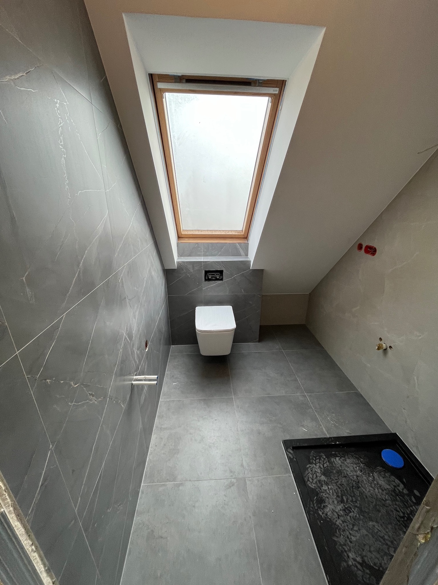 Wnętrze łazienki na poddaszu z szarymi płytkami imitującymi marmur, wiszącą toaletą, oknem dachowym i brodzikiem w trakcie montażu.