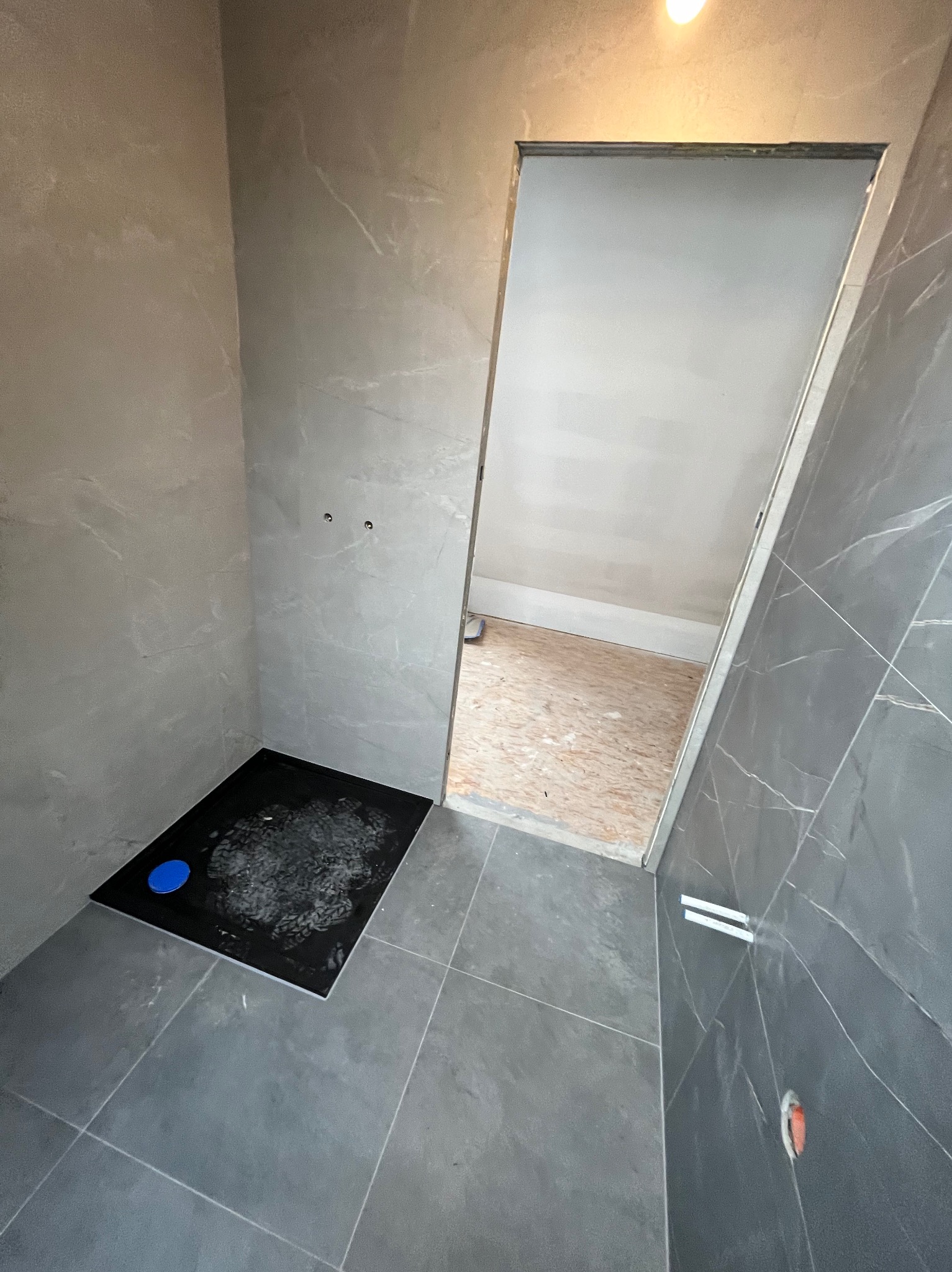 Wnętrze łazienki w trakcie wykańczania, widoczny czarny brodzik z niebieskim odpływem, szare płytki imitujące marmur na ścianach i podłodze, surowy otwór drzwiowy z widoczną podłogą z płyt OSB...