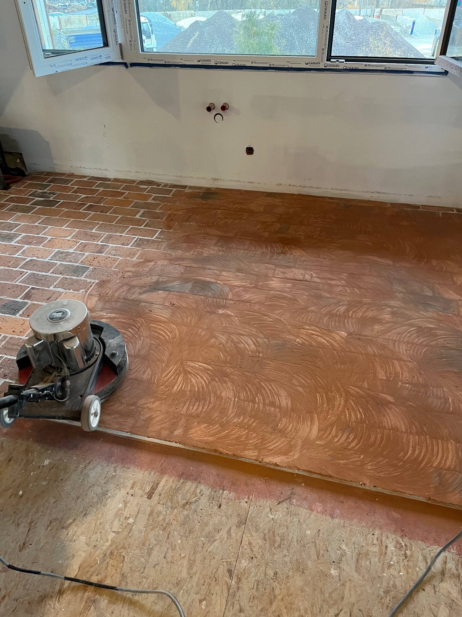 Szlifowanie ceglanej podłogi w pomieszczeniu poddaszowym; widoczna maszyna szlifująca, częściowo wyszlifowana powierzchnia, okno z widokiem na plac budowy.