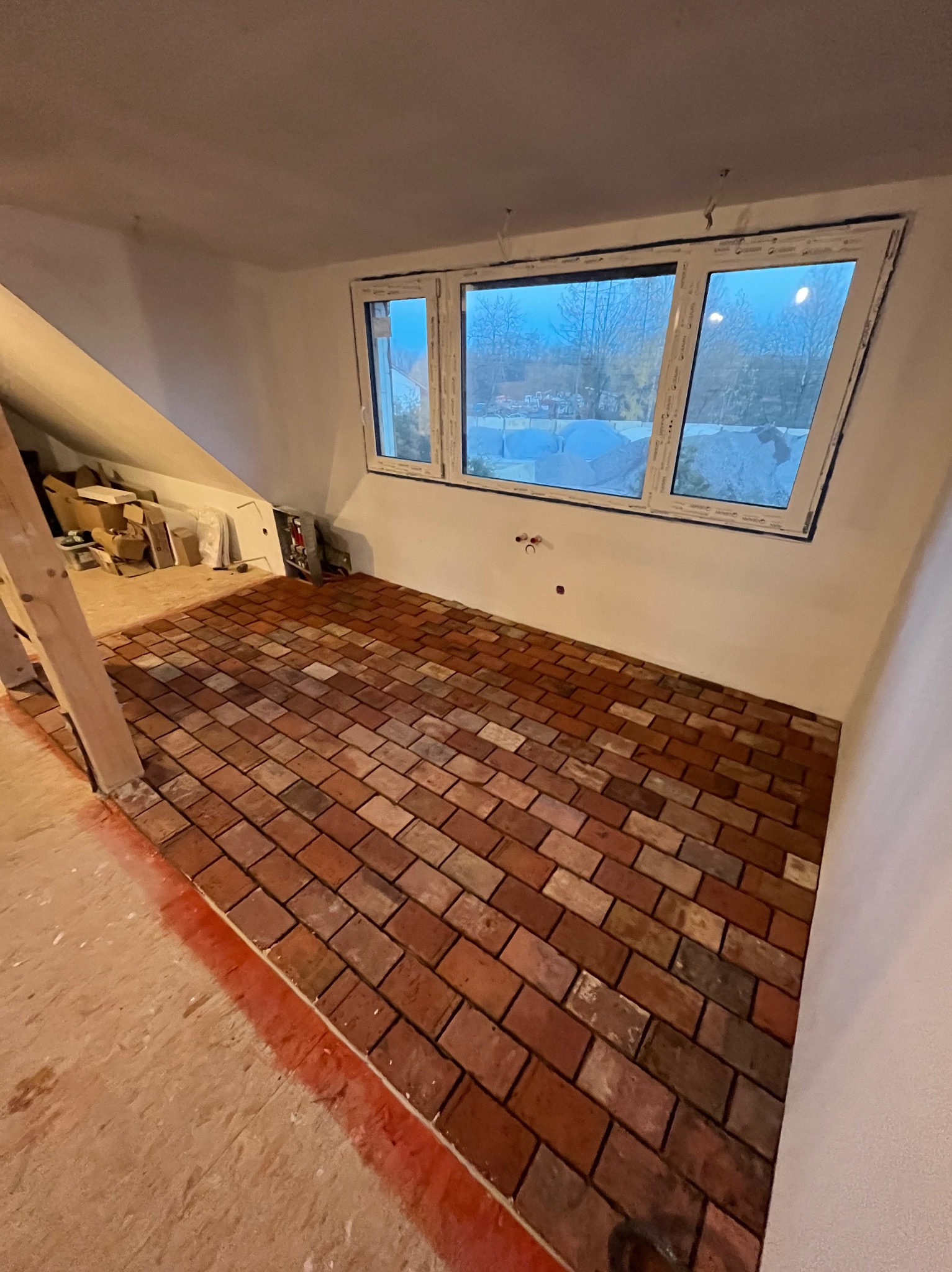 Nowo ułożona ceglana podłoga w pomieszczeniu ze skosami, widoczne okno i przygotowane ściany, częściowo zakryta drewniana belka.