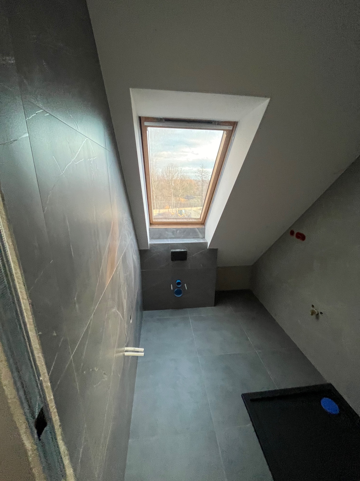 Szara łazienka na poddaszu z oknem dachowym, widoczne płytki imitujące marmur i czarny brodzik z niebieską naklejką, instalacja podtynkową i wystające rurki.