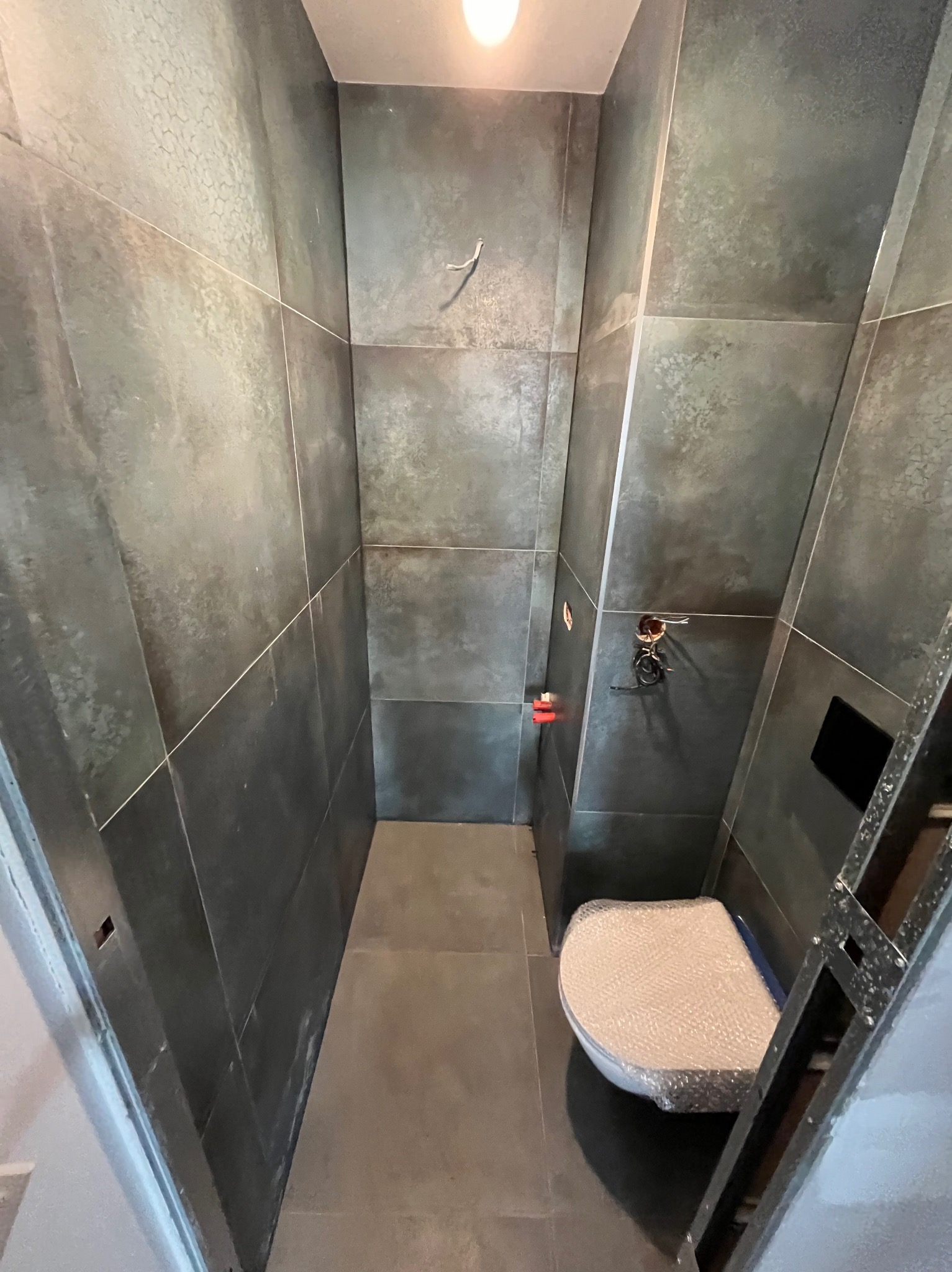 Wąska łazienka w trakcie wykańczania, ciemne płytki na ścianach, podłoga z szarych płytek, częściowo zamontowana toaleta zabezpieczona folią bąbelkową, widoczne przewody elektryczne i fragment...