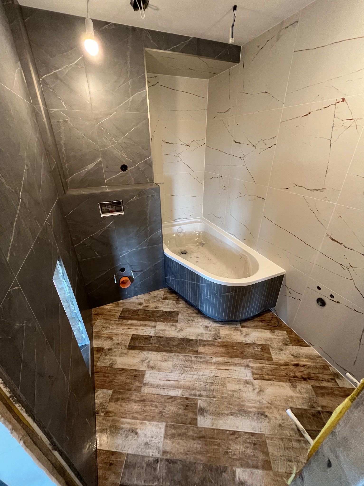 Wnętrze łazienki w trakcie prac wykończeniowych: częściowo ułożone płytki imitujące marmur na ścianach, drewnopodobna podłoga, zamontowana wanna z obudową w odcieniach szarości oraz zabudowa...