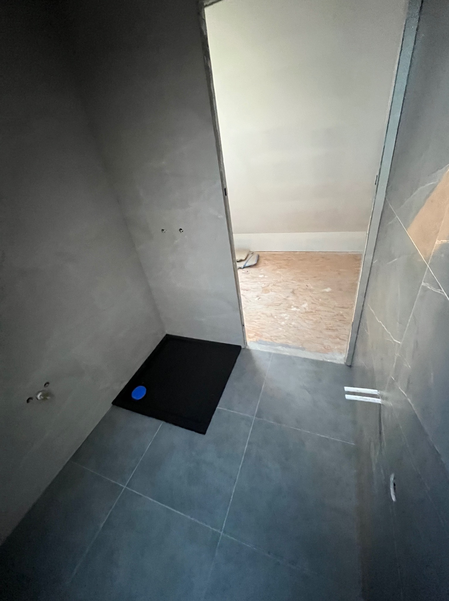 Niedokończona łazienka z czarnym brodzikiem i surowymi szarymi ścianami, widoczne przygotowania do montażu armatury, podłoga wyłożona szarą płytką, widok na sąsiednie pomieszczenie z surową podłogą.