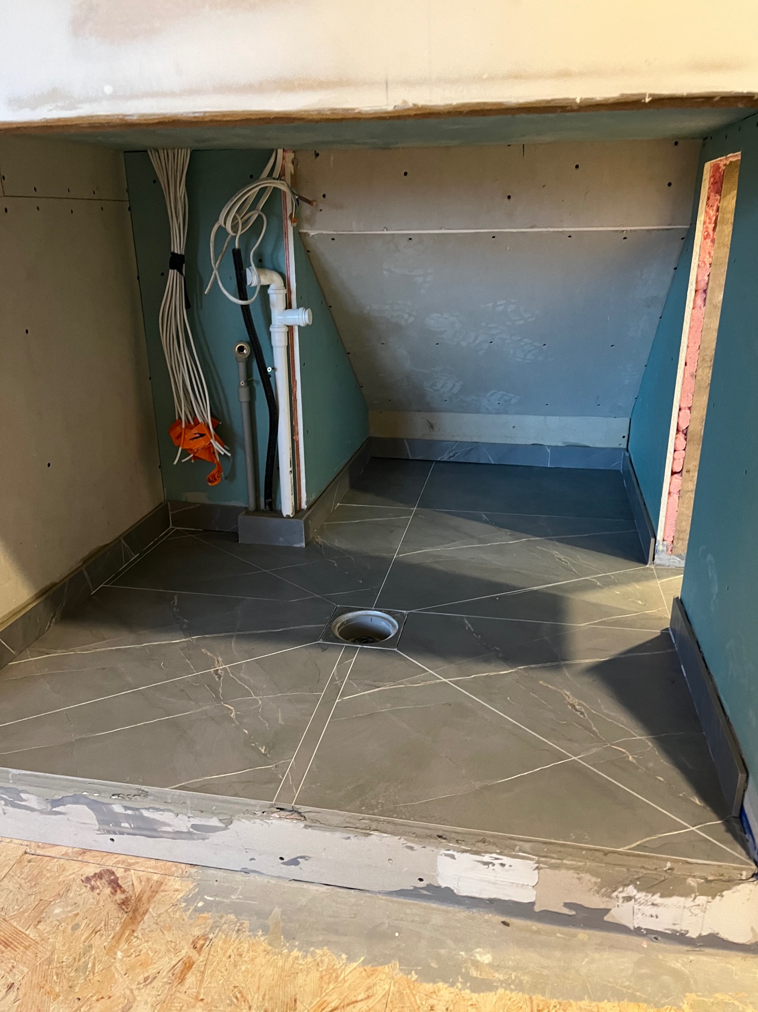 Niedokończona kabina prysznicowa z szarą marmurową podłogą i odpływem liniowym, widoczne kable i instalacja wodna, ściany w surowym stanie przed wykończeniem.