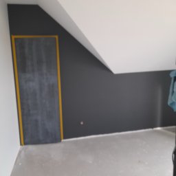 Darson Remonty Dariusz Kukulski - Pokój na poddaszu z pomalowaną na szaro ścianą i ościeżnicą zabezpieczoną żółtą taśmą malarską, surowa podłoga przed wykończeniem.
