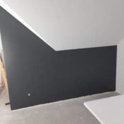 Darson Remonty Dariusz Kukulski - Ściana pomalowana na grafitowo z widocznym otworem na gniazdko elektryczne, biały sufit pod skosem, fragment drabiny malarskiej w lewym rogu i blat roboczy w prawym dolnym rogu.