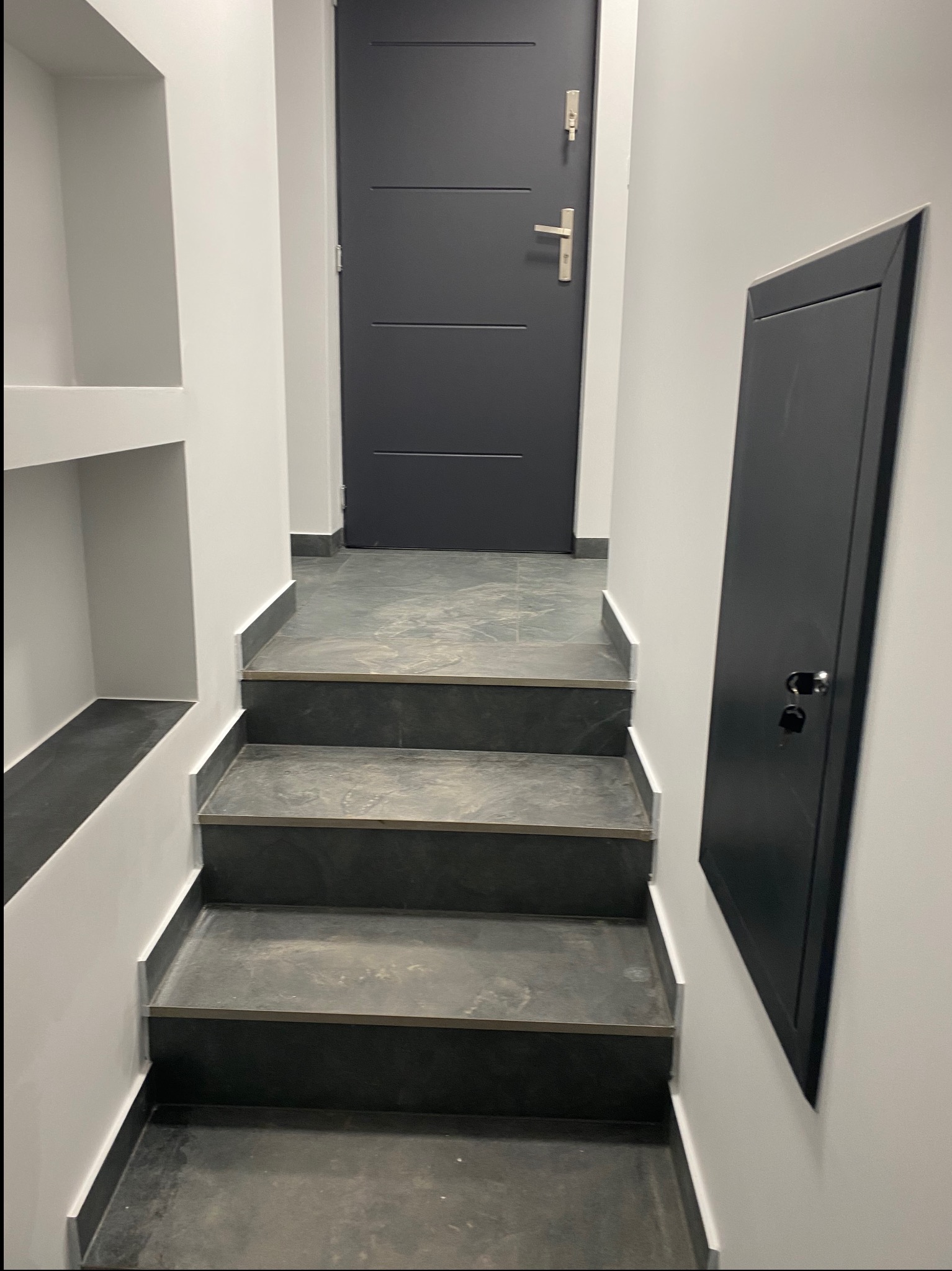Klatka schodowa z trzema stopniami wykończonymi szarym kamieniem, białe ściany z wnękami, szara drzwi wejściowe i czarna szafka rewizyjna.