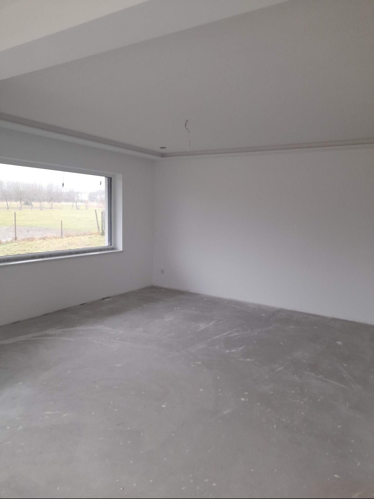 Pusty pokój z gładkimi, białymi ścianami i szarym betonowym podłożem, widok na zewnątrz przez okno z widocznym krajobrazem polnym.