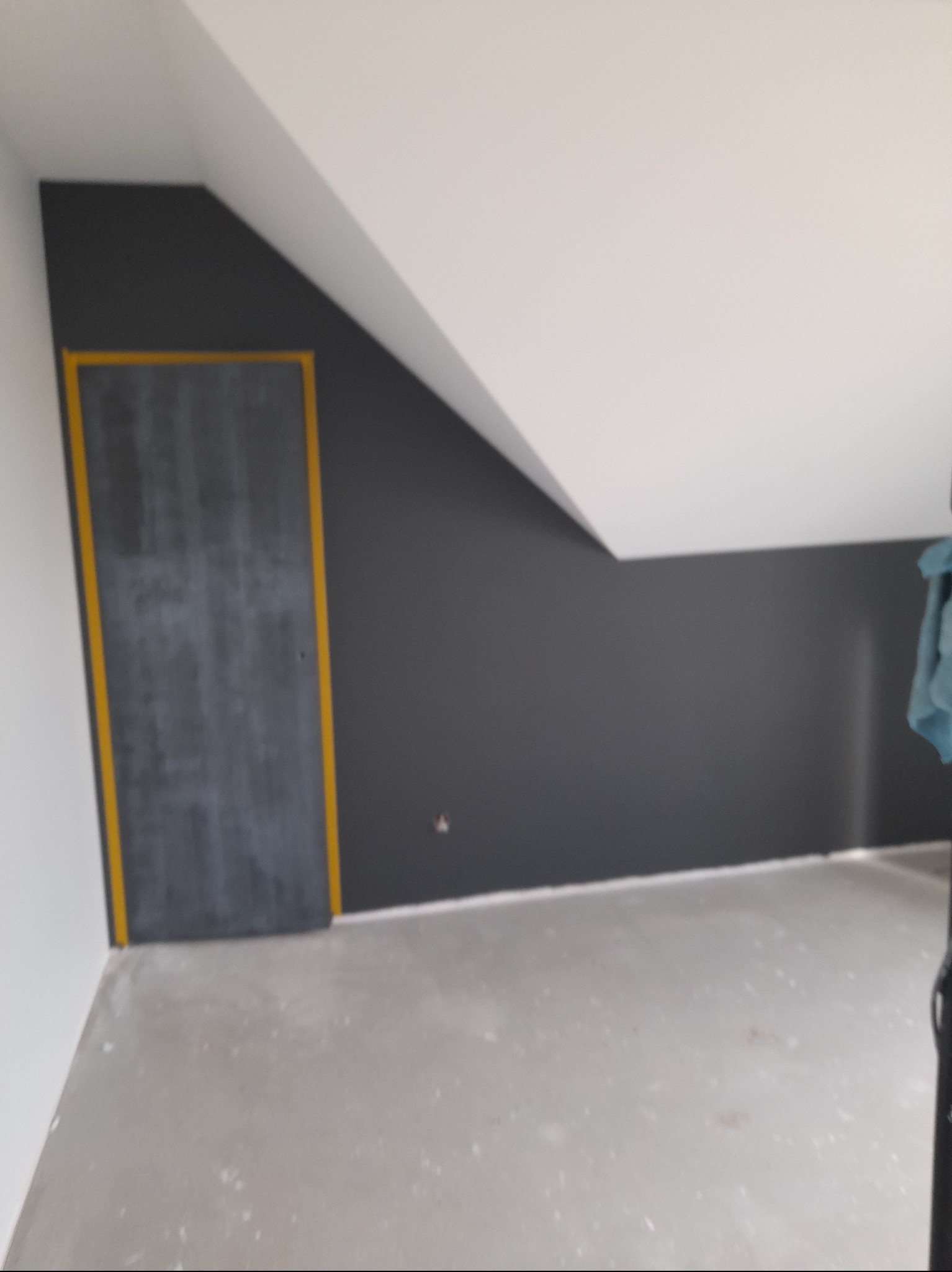 Pokój na poddaszu z pomalowaną na szaro ścianą i ościeżnicą zabezpieczoną żółtą taśmą malarską, surowa podłoga przed wykończeniem.