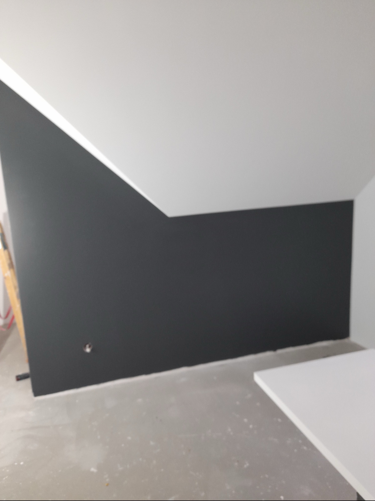 Ściana pomalowana na grafitowo z widocznym otworem na gniazdko elektryczne, biały sufit pod skosem, fragment drabiny malarskiej w lewym rogu i blat roboczy w prawym dolnym rogu.
