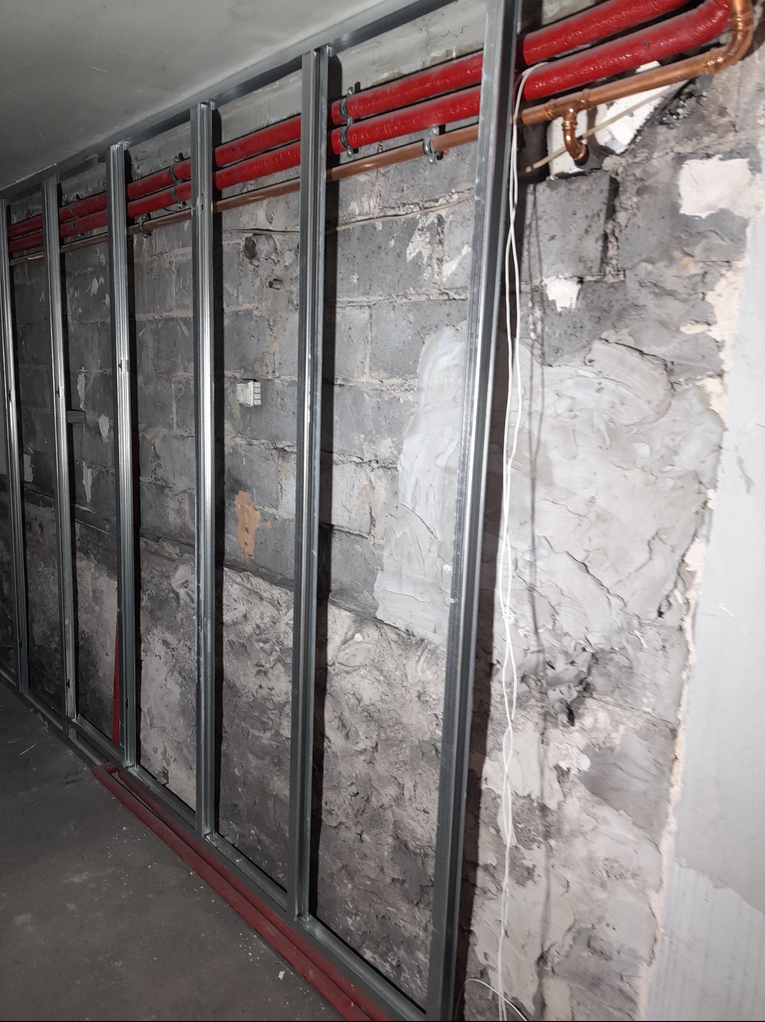 Stelaż z profili metalowych przy nierównej, częściowo otynkowanej ścianie z instalacją wodną z rur miedzianych i czerwonych rur PCV.