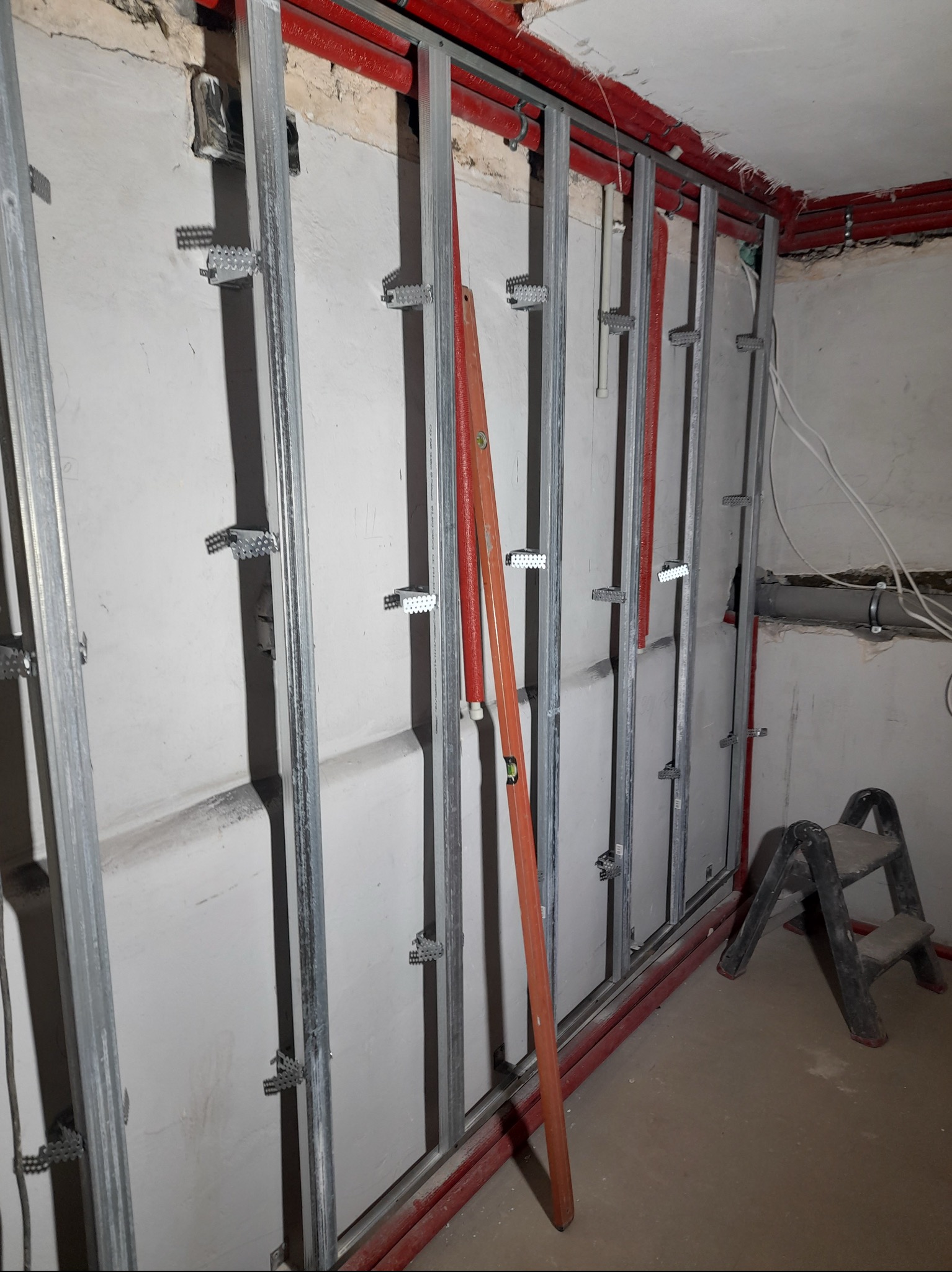 Stelaż z profili metalowych przymocowany do ściany za pomocą uchwytów, pionowy poziom w kolorze pomarańczowym oparty o konstrukcję, widoczne instalacje w czerwonej osłonie i drabinka.