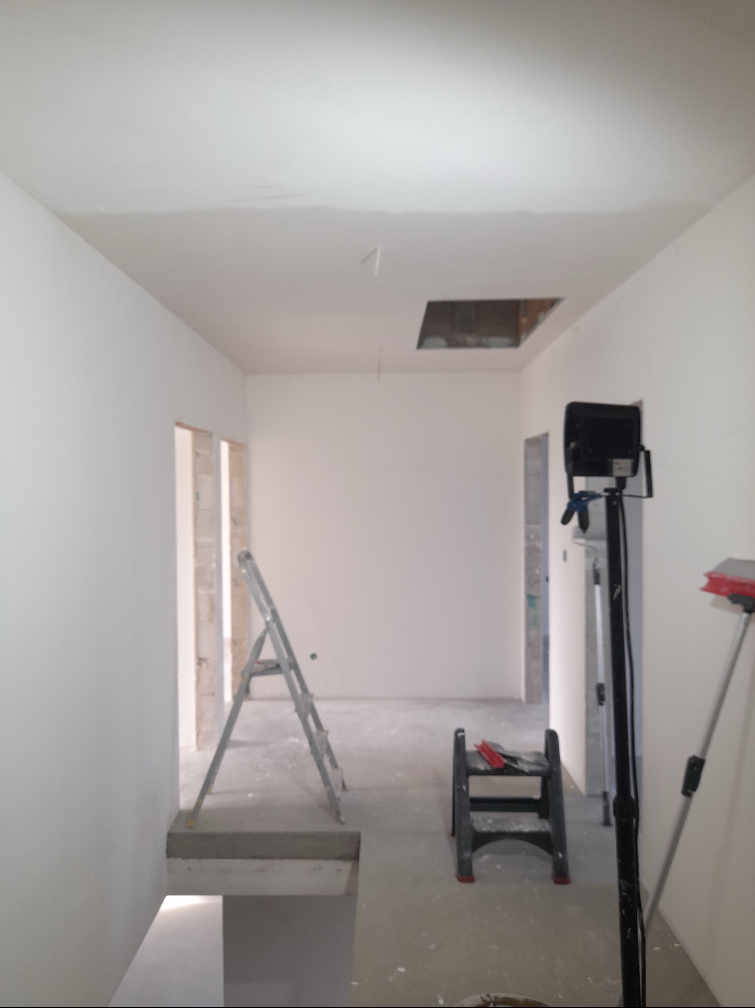 Wnętrze w trakcie remontu: białe ściany, drabina aluminiowa, otwór w suficie, lampa halogenowa na statywie, mały stołek z narzędziami.