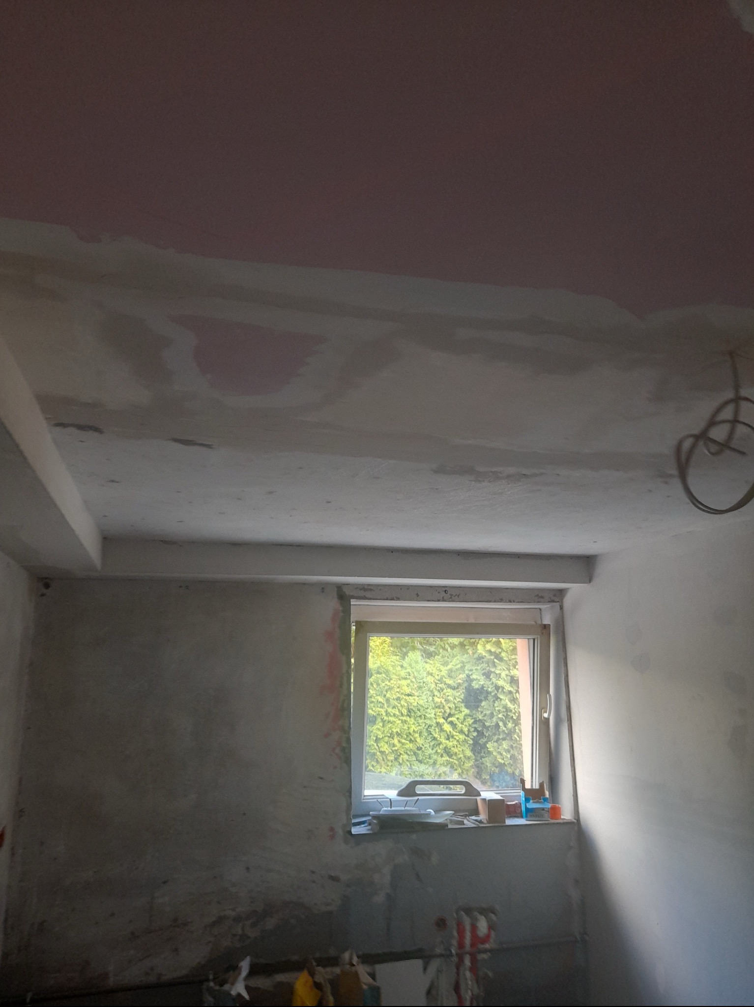 Pokój w trakcie remontu: częściowo wyszpachlowany sufit z widocznymi śladami po łatach, ściany z surowym tynkiem, okno z widokiem na zieleń, kable zwisające z sufitu.