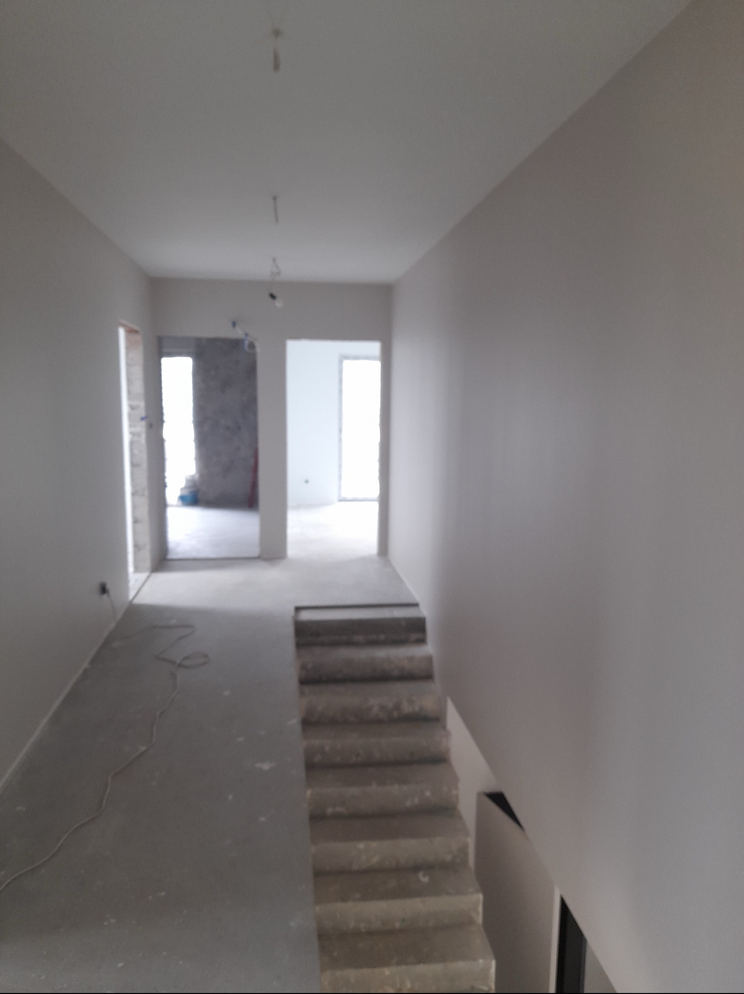 Wnętrze domu w trakcie remontu, widoczne gładkie, szare ściany, surowe schody betonowe i kable na podłodze.