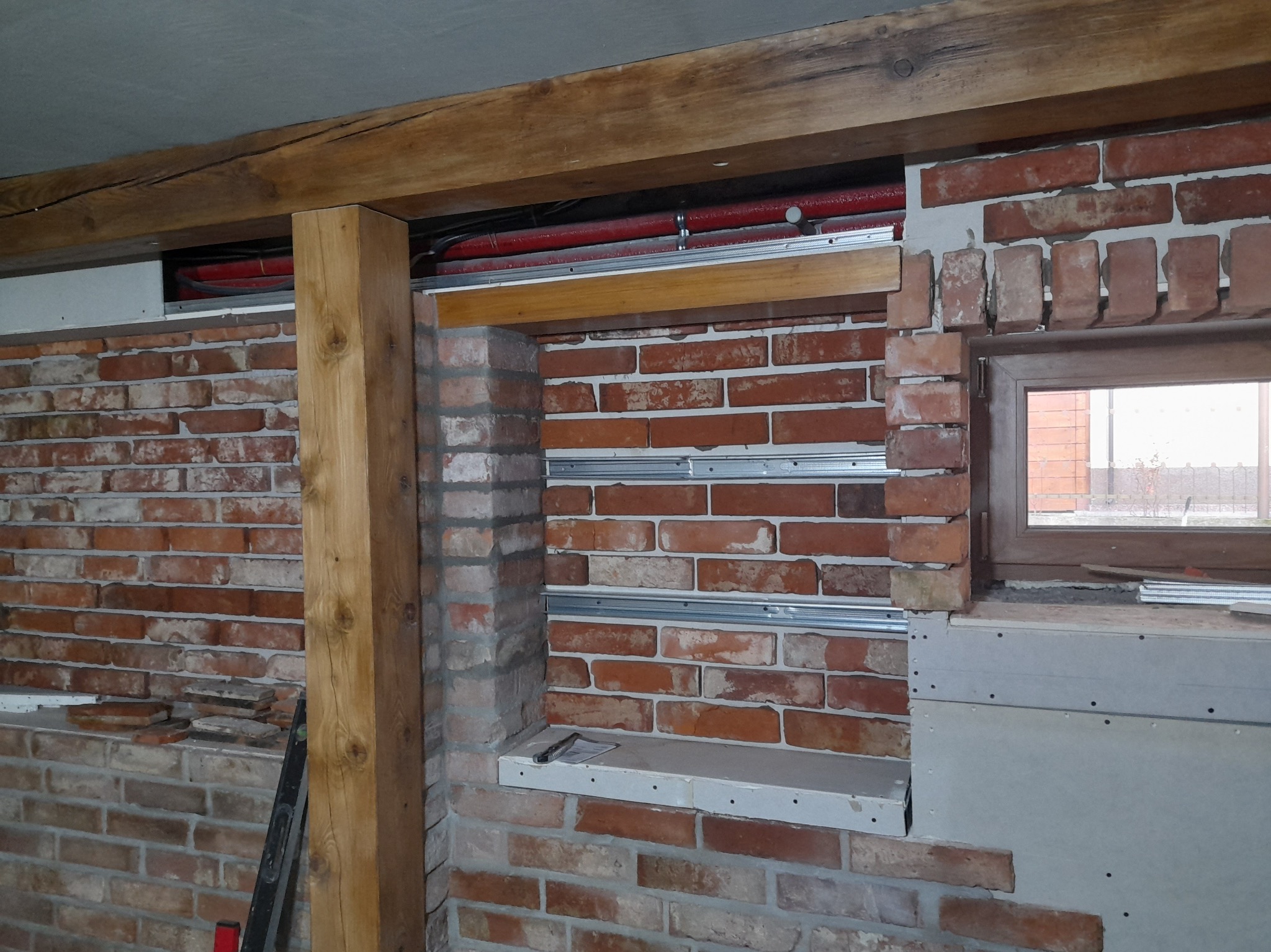 Wnętrze w trakcie remontu: fragment ściany z cegły z drewnianą belką i słupem, zamontowane profile do płyt karton-gips, widoczne rury instalacyjne.