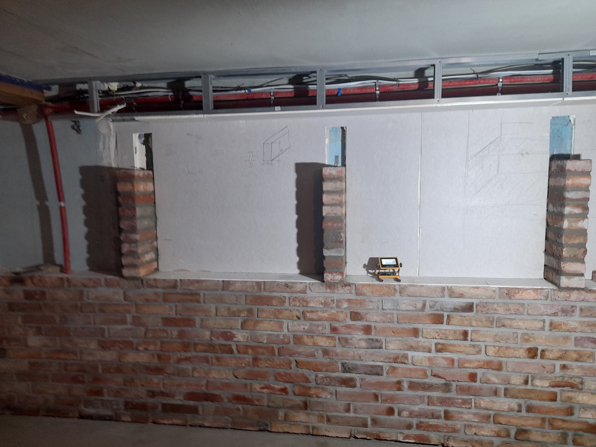 Ceglana ściana z fragmentami płyty karton gips, widoczne metalowe profile pod sufitem i laserowy niwelator na murku.