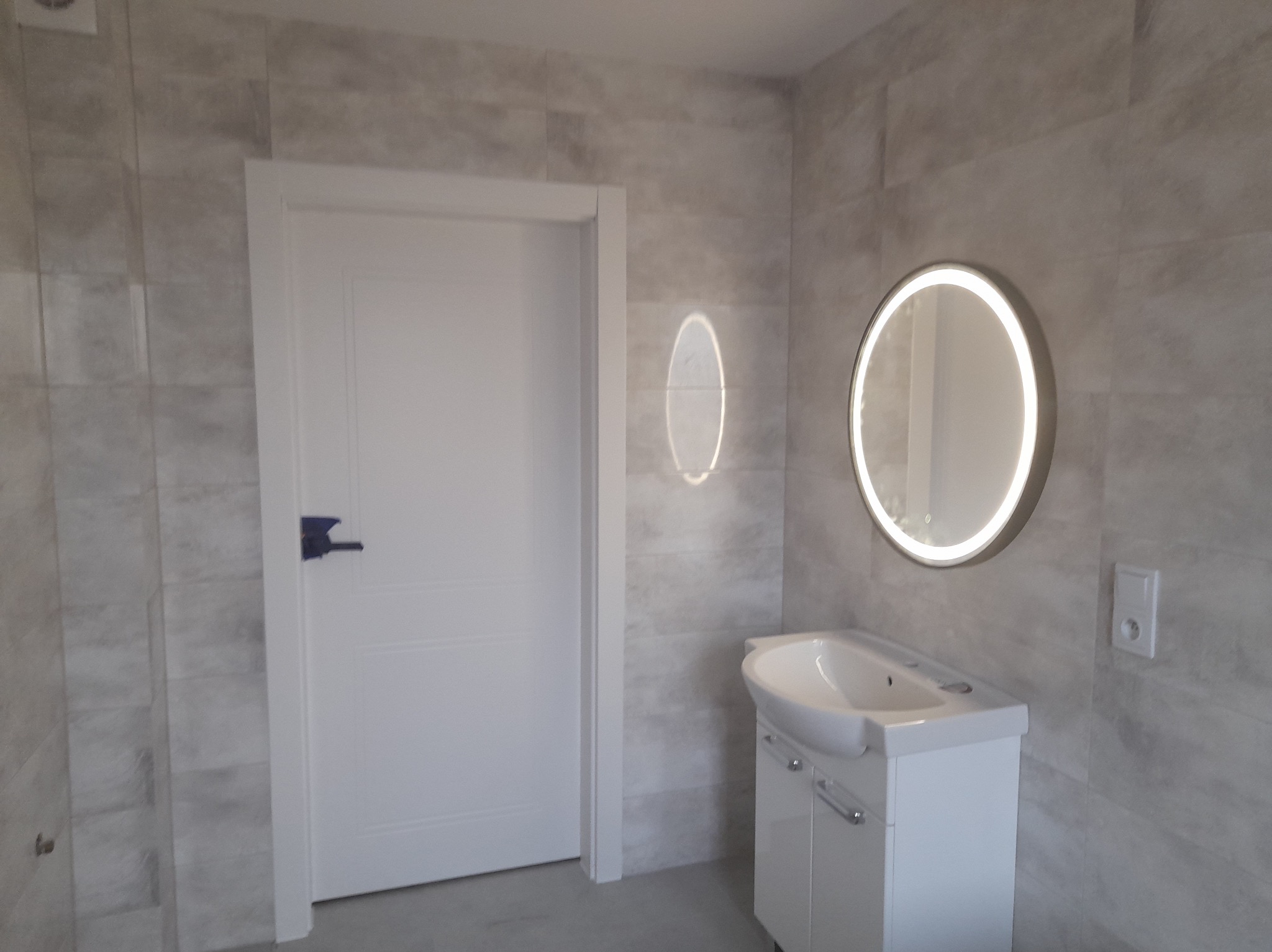 Wykończona łazienka z szarymi płytkami imitującymi kamień, białą umywalką z szafką, okrągłym lustrem z podświetleniem LED i białą, prostą, zamkniętą drzwiami z czarną klamką.