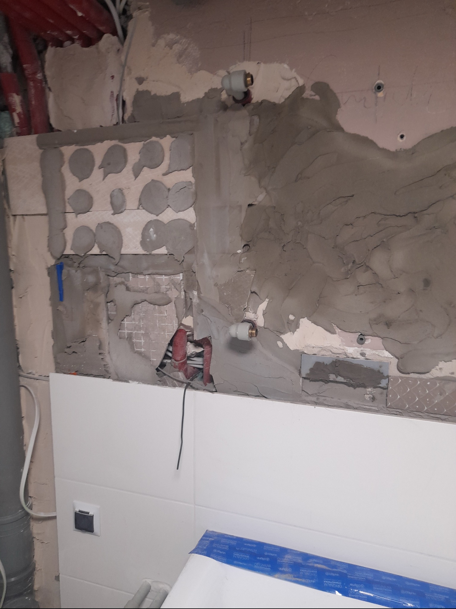 Ściana w trakcie remontu: widoczne fragmenty starej i nowej glazury, instalacja wodna, ślady po szpachlowaniu i przygotowaniu podłoża pod płytki.
