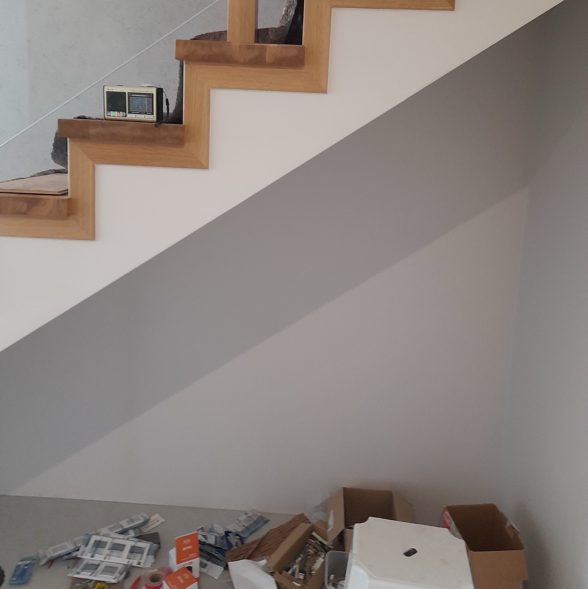 Ściana pomalowana na szaro pod drewnianymi schodami z białą balustradą, na stopniu stoi radio, a na podłodze porozrzucane materiały budowlane i kartony.