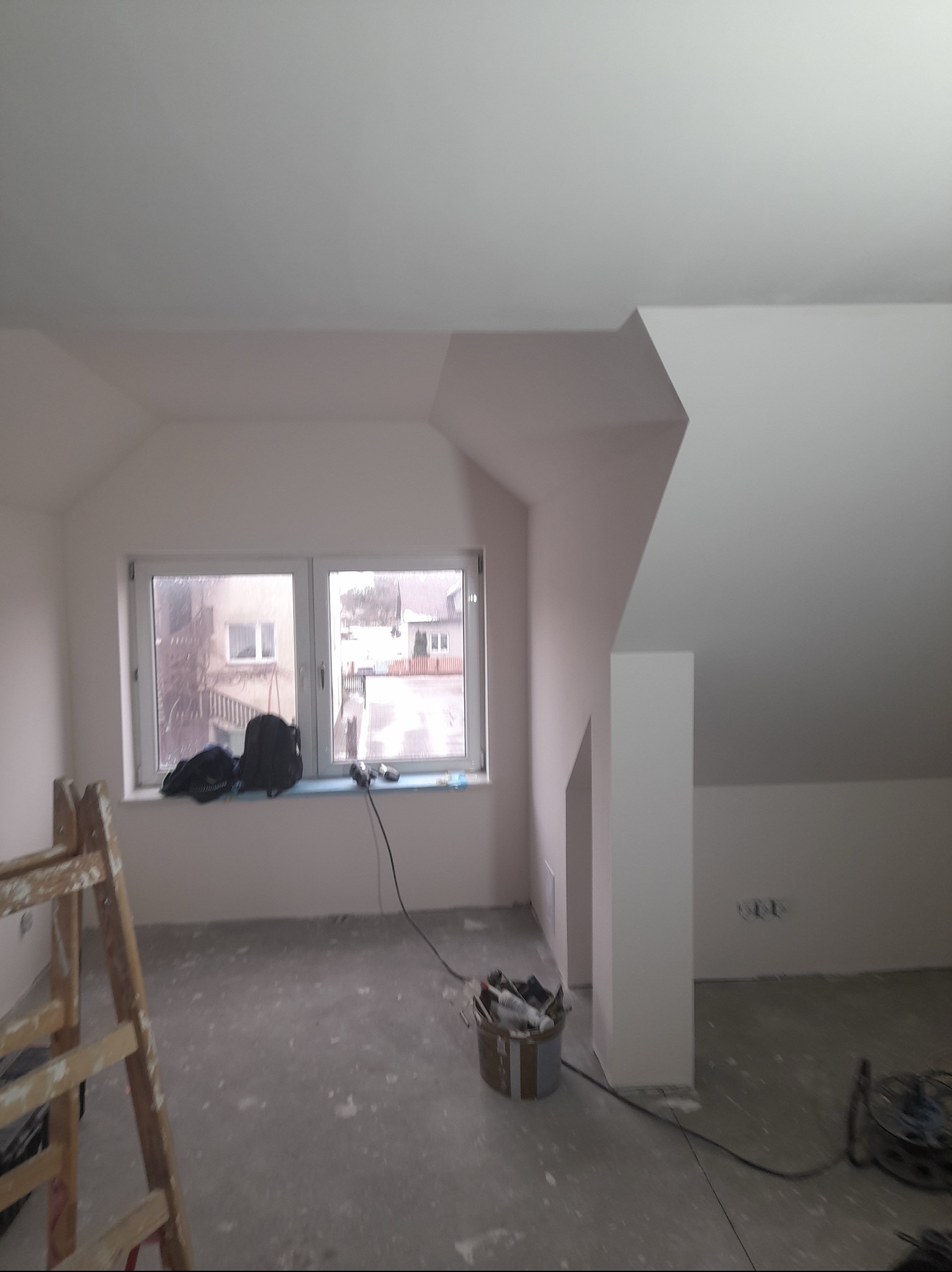 Poddasze w trakcie prac wykończeniowych, ściany i sufit po nałożeniu gładzi, widoczne narzędzia i drabina malarska, okno z widokiem na zabudowę jednorodzinną.