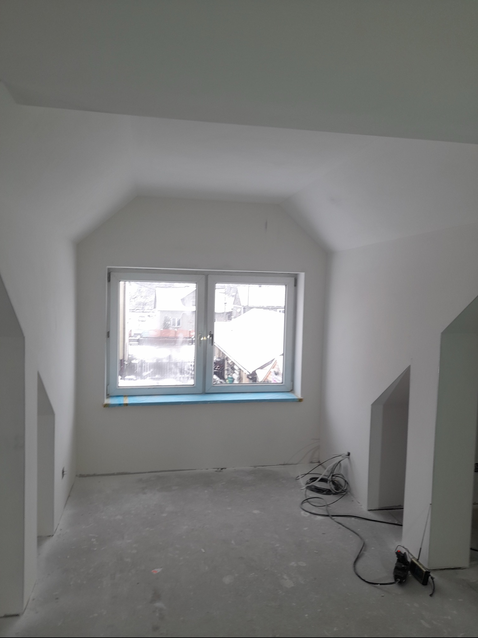 Poddasze po remoncie, świeżo pomalowane na biało, z oknem zabezpieczonym folią malarską, widoczne przewody elektryczne na podłodze.