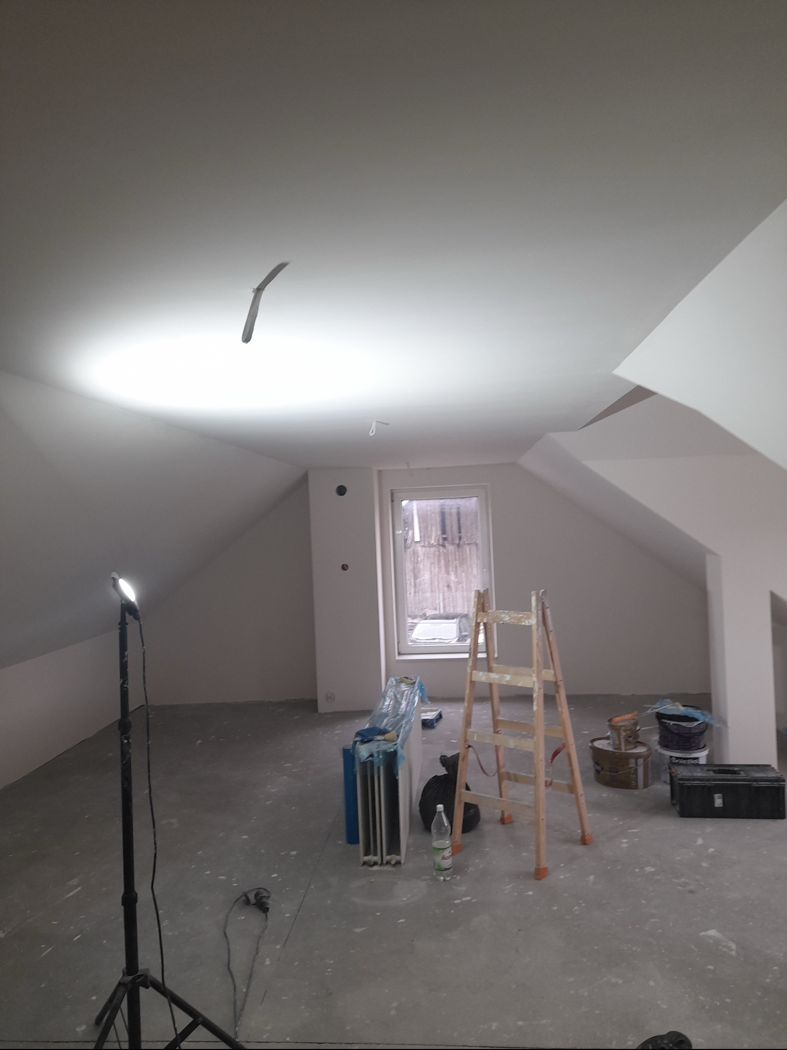 Poddasze po remoncie, świeżo pomalowane na biało, widoczna drabina, lampa stojąca, wiadro z farbą i narzędzia malarskie.