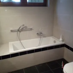 Wykończona łazienka z białą wanną obudowaną płytkami z czarnym pasem, szara glazura na ścianach, ciemna podłoga z płytek, okno z widokiem na iglaki.