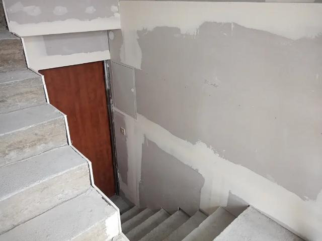 Klatka schodowa w trakcie prac wykończeniowych: surowe betonowe schody, częściowo zagruntowane ściany z widocznymi śladami szpachlowania, brązowe drzwi.