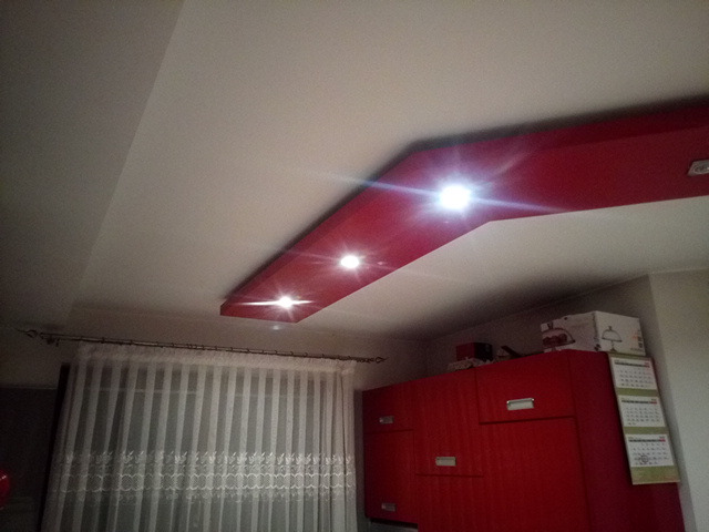 Wykończony sufit podwieszany z czerwonym elementem i wbudowanym oświetleniem punktowym w pokoju z zasłonami i czerwonymi szafkami.