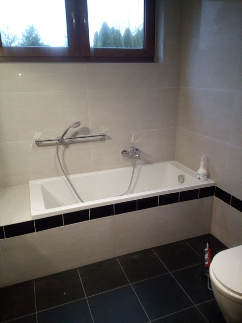 Wykończona łazienka z białą wanną obudowaną płytkami z czarnym pasem, szara glazura na ścianach, ciemna podłoga z płytek, okno z widokiem na iglaki.