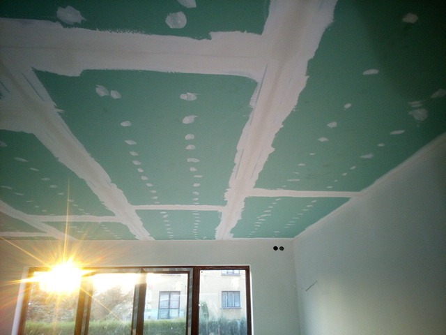 Płyty gipsowo-kartonowe na suficie, szpachlowane łączenia, widok na okno z promieniami słonecznymi.