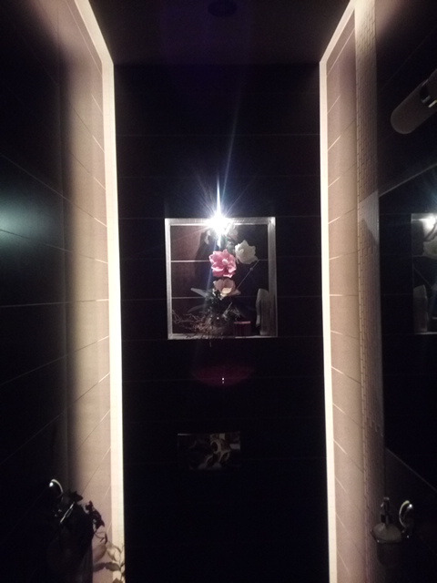 Ciemna łazienka z czarnymi płytkami, oświetlona pionowymi pasami światła po bokach i odbiciem światła w lustrze z kwiatami.