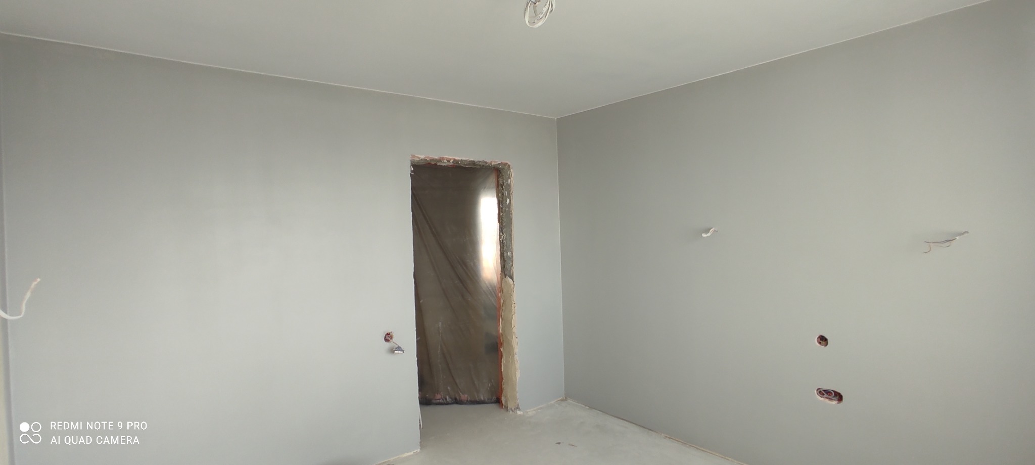 Pokój po świeżym pomalowaniu na szaro, widoczne wystające kable ze ściany i sufitu, oraz surowy otwór drzwiowy.