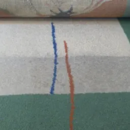 Fragment zwiniętego dywanu z geometrycznym wzorem (biały trapez z niebieską i pomarańczową linią) na zielonym tle, oferowany w Zielonej Górze.