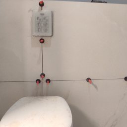 Bud Vas Vasyl Floreskul - Świeżo ułożone, duże płytki ceramiczne na ścianie w łazience z widocznymi systemami poziomowania płytek i częściowo widoczną toaletą w dolnej części kadru.