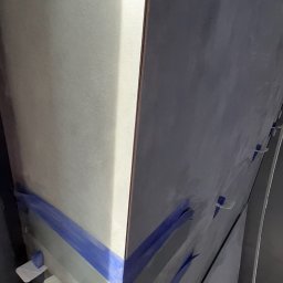 Bud Vas Vasyl Floreskul - Układanie szarych płytek na narożniku ściany, zabezpieczonych niebieską taśmą i białymi krzyżykami dystansowymi, widoczne narzędzia i wiadro z gąbkami.