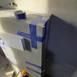 Bud Vas Vasyl Floreskul - Szara ściana w trakcie układania płytek, zabezpieczona niebieską taśmą malarską i klinami dystansowymi, widoczny fragment wentylacji, ujęcie z dołu.