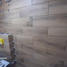 Bud Vas Vasyl Floreskul - Ściana w trakcie wykańczania z ułożonymi płytkami imitującymi drewno, widoczne narzędzia glazurnicze i plastikowe krzyżyki dystansowe.